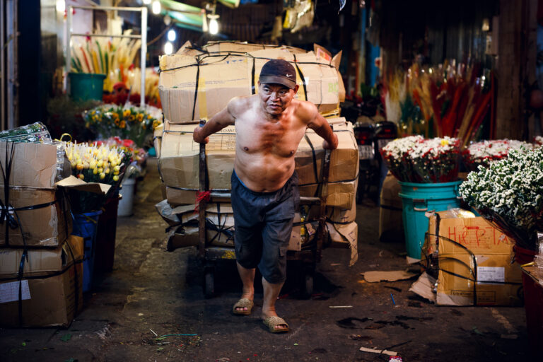 A Vietnamese man is captured in a candid portrait pulling a cart full of boxes at a flower market in Ho Chi Minh