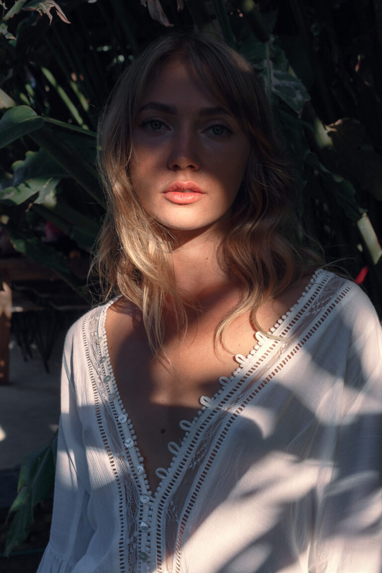 A young woman stands in the shadows of a bush wearing a white dress whilst posing for a portrait