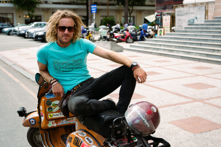 a young man poses for a portrait whilst sitting with his legs up on an orange scooter