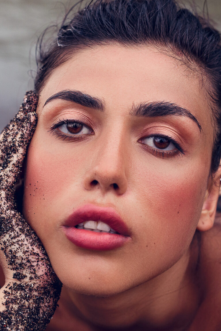 A young woman holds her head in her black sand covered hand whilst she poses for a portrait