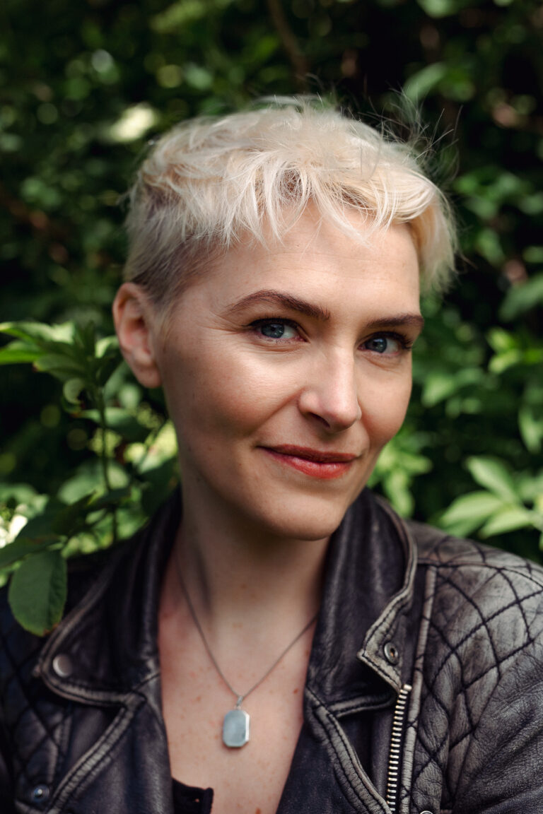 An outdoor headshot of a young smiling woman wearing a leather jacket