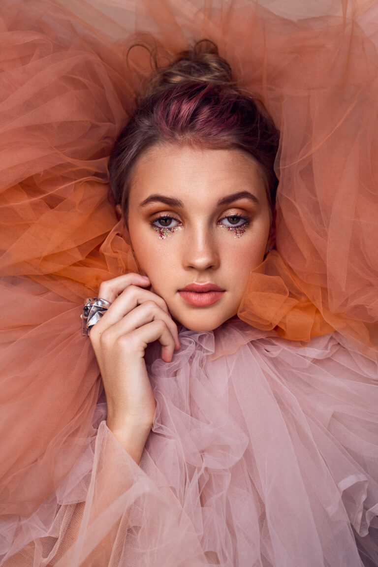 A young woman with her hand next to her face surrounded by pink tulle poses during a fashion shoot