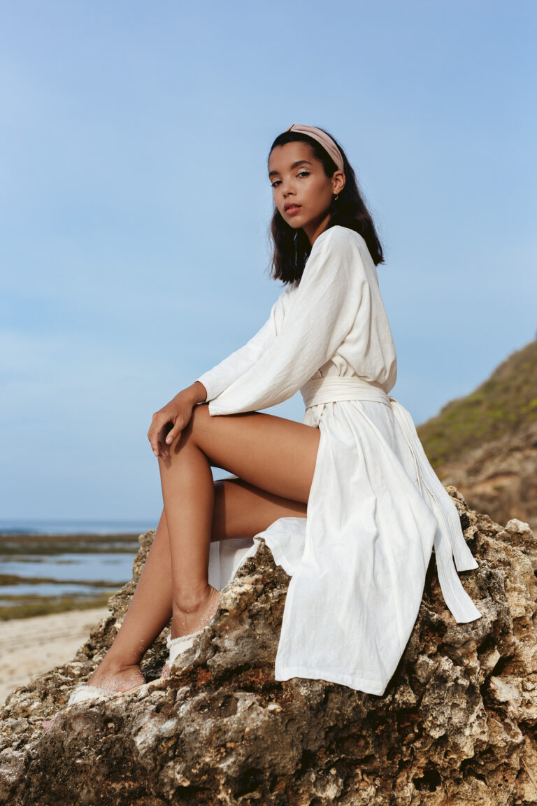 A young woman poses for a fashion portrait wearing a white dress sitting on a rock on a beach in Bali