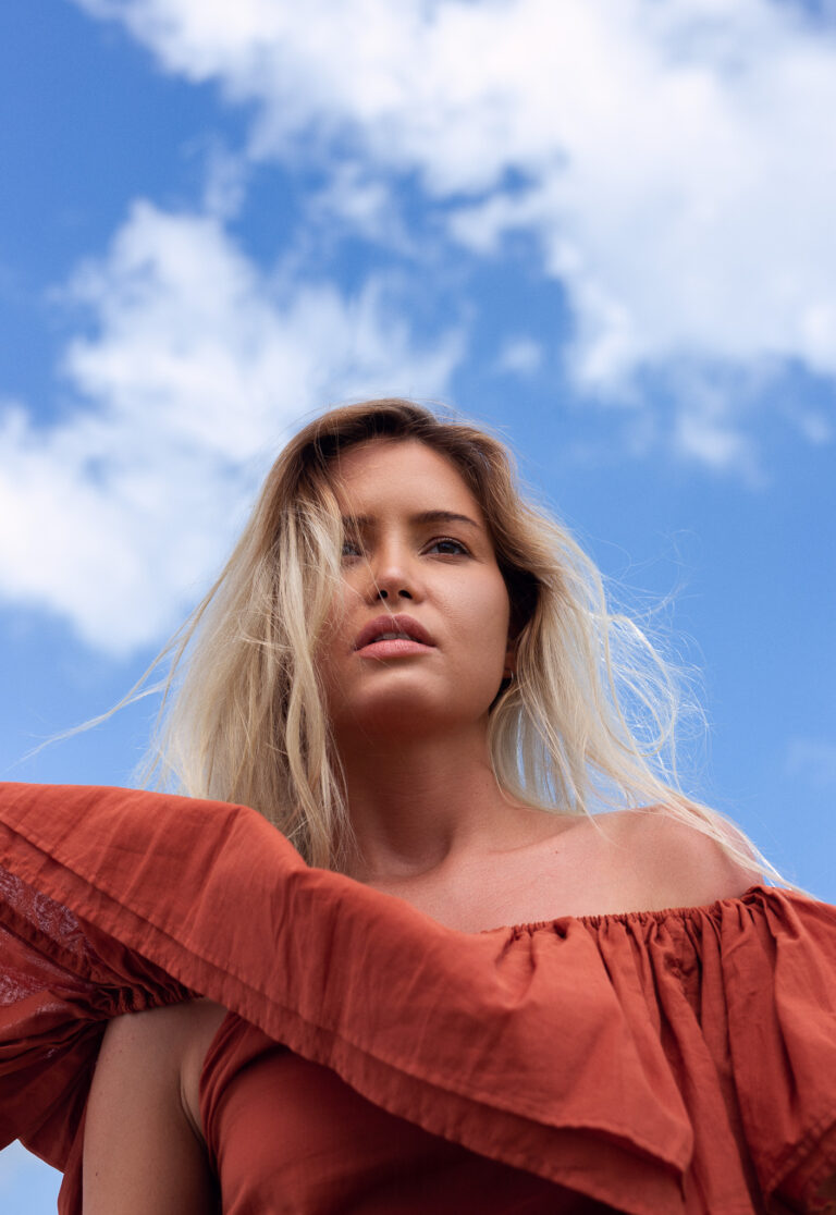A young blonde woman wearing a red top with sky and clouds behind her poses during a fashion shoot