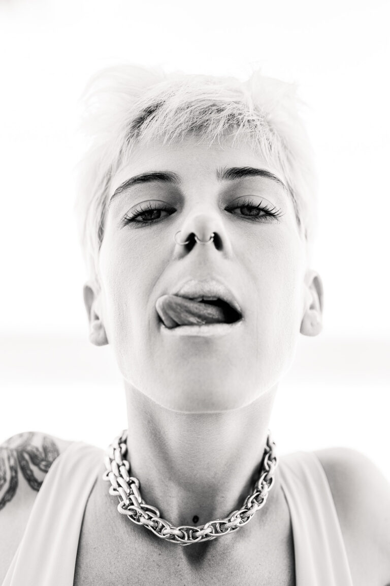 An edgy woman with short blonde hair and nose piercings licks her lips while posing for a fashion portrait
