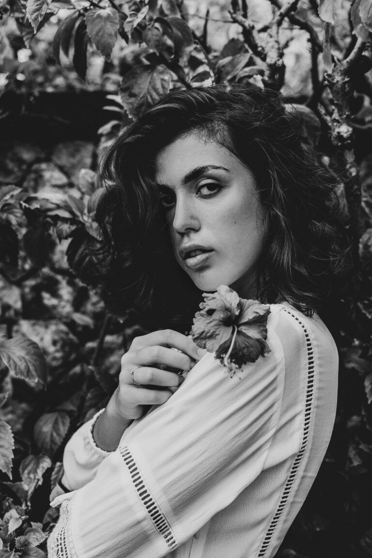 A young woman in a white dress holding a flower up to her shoulder poses for a portrait