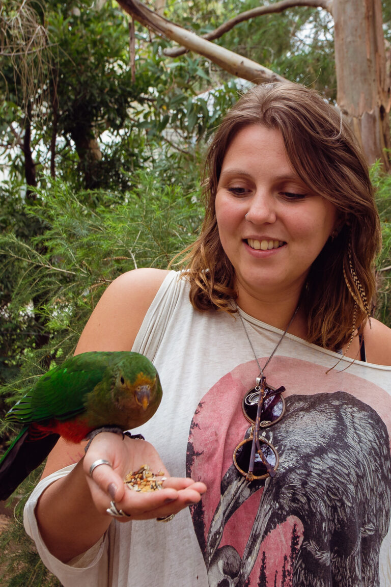 Olga Rozewin smiling and holding bird feed in her palm with a green parrot sitting on her hand