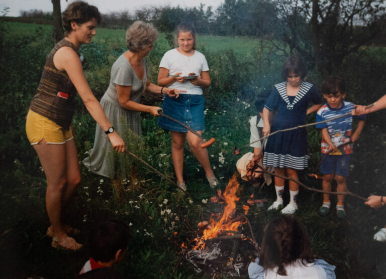 family of varying ages standing around a campfire cooking sausages on sticks