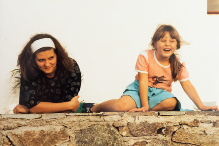 a young woman standing leaning on a wall with a young female child sitting on the wall and laughing