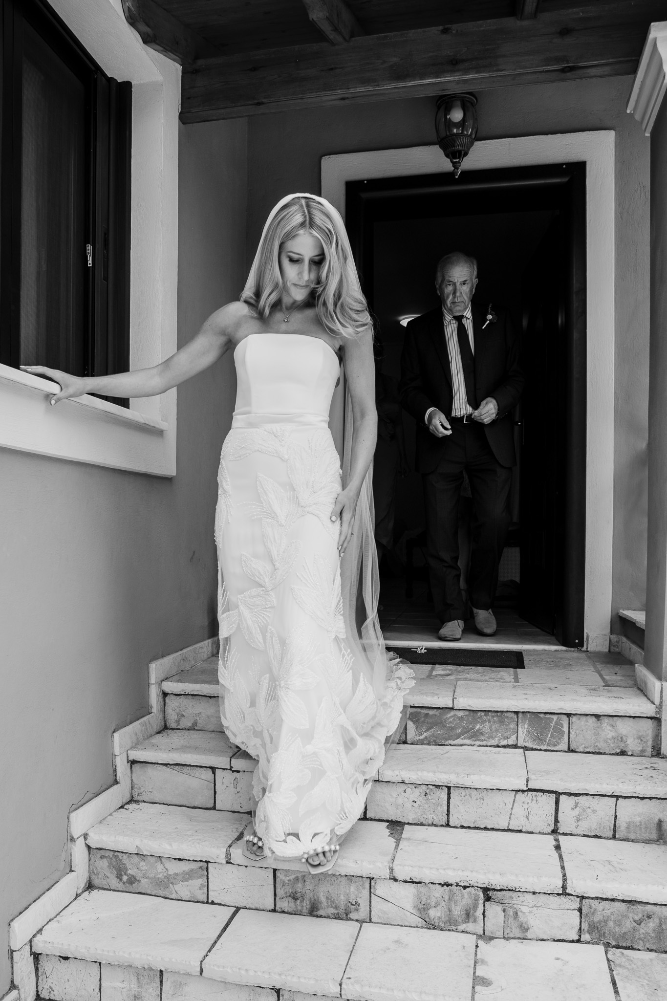 Alice walks down the stairs in her wedding dress with her dad at her villa in Crete.