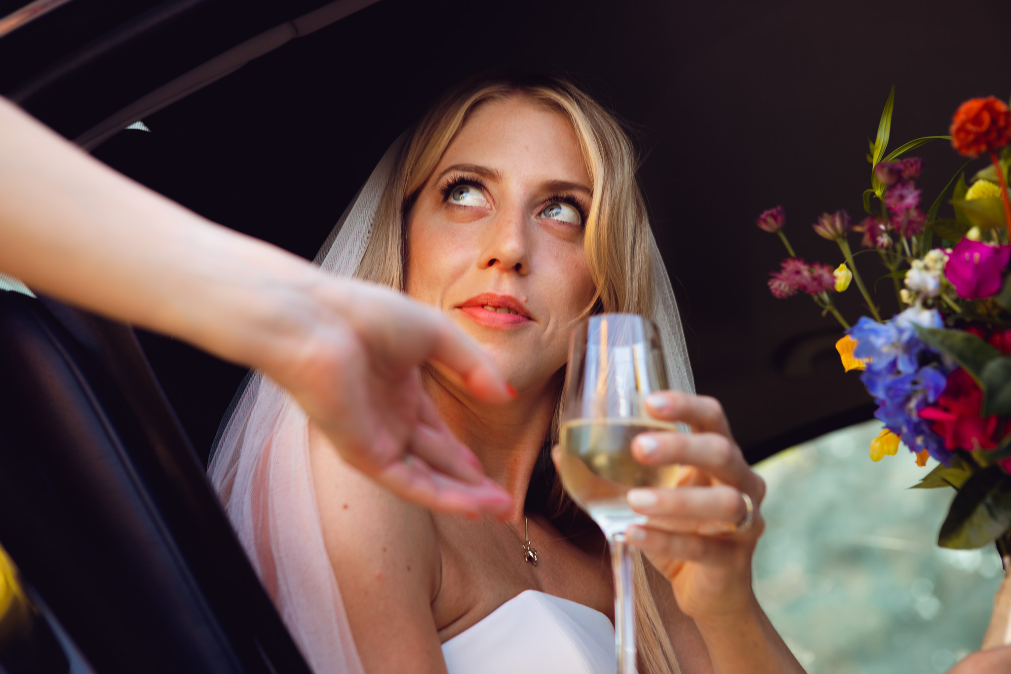 Bride sits in car and smiles whilst someone hands her a champagne flute