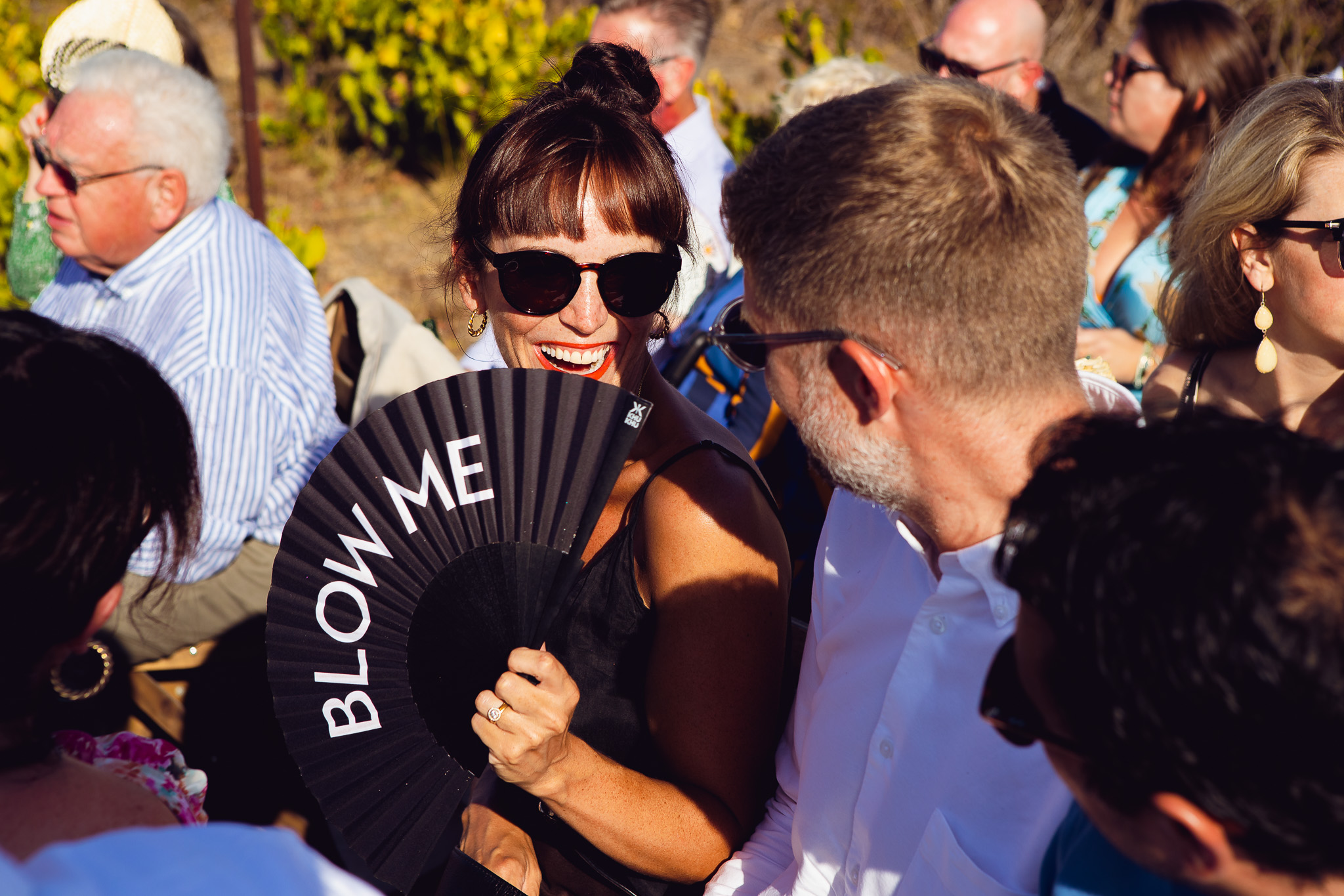 Guest smiles holding a blow me fan waiting for the bride to arrive at a wedding ceremony in Ambelonas Vineyard, Corfu