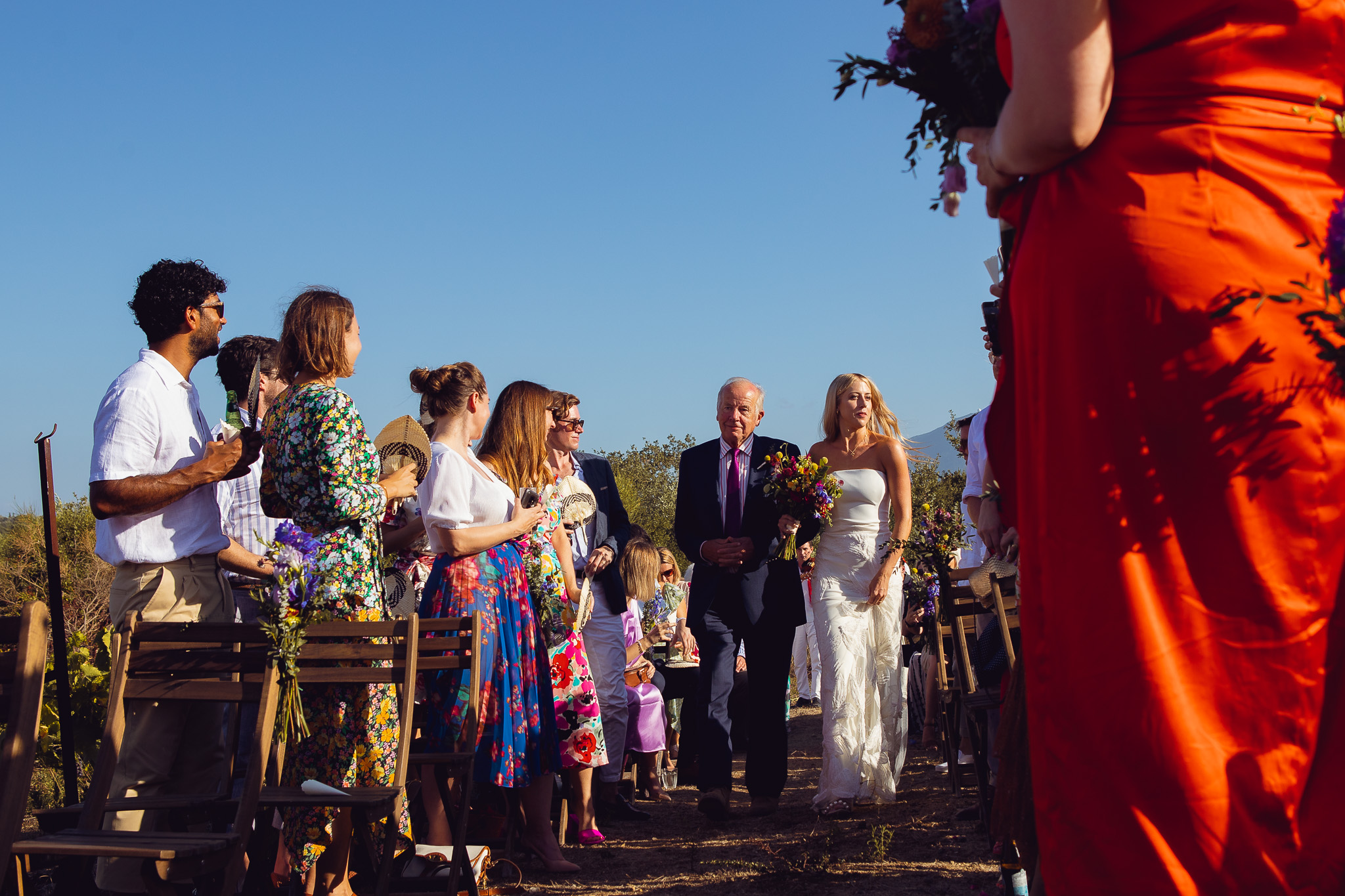 Guests stand whilst bride walks down aisle with her dad at a wedding ceremony in Ambelonas Vineyard, Corfu