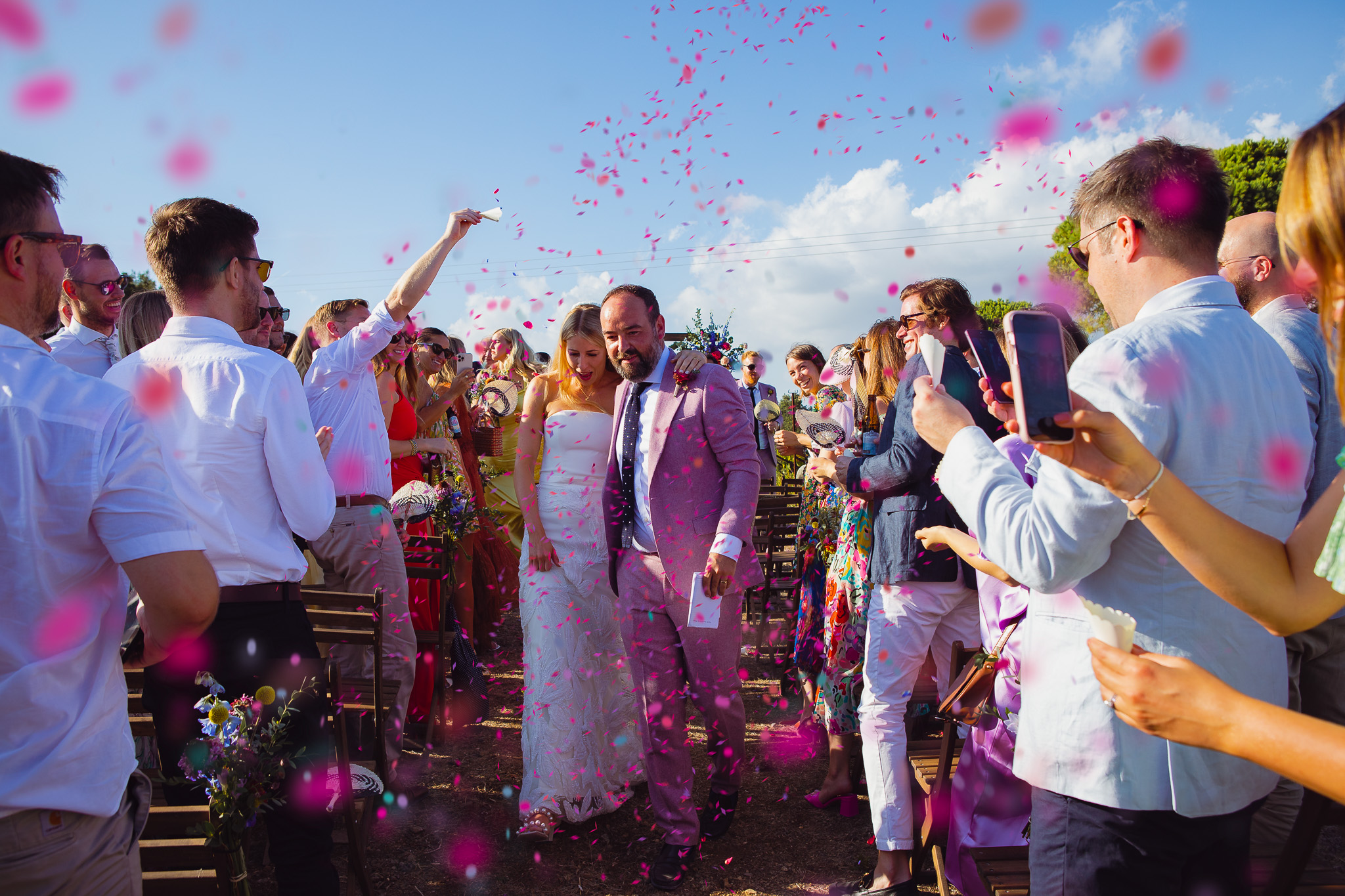Newlywed bride and groom walk through their guests as they throw pink confetti after their wedding ceremony