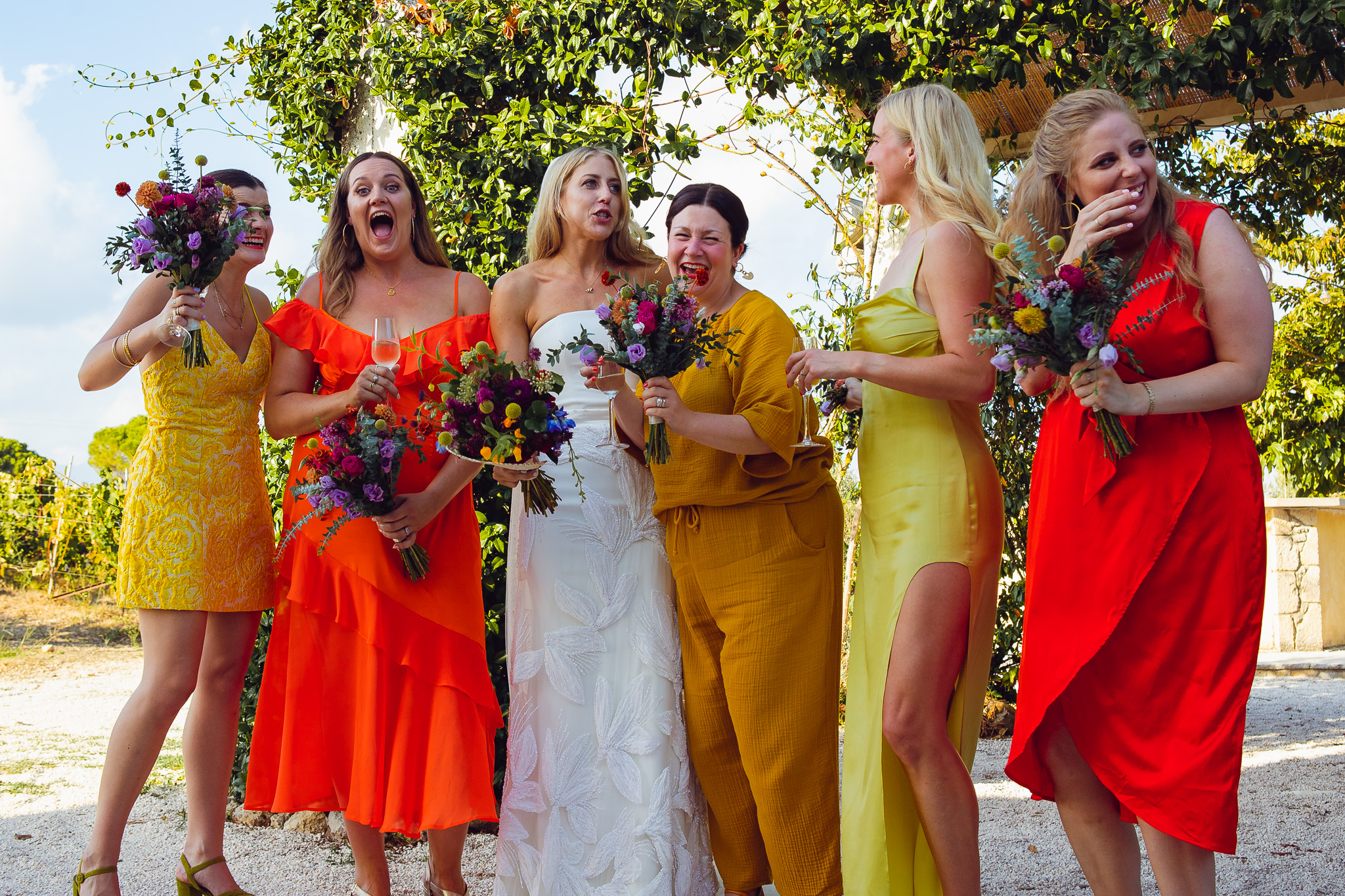 The bride and five brightly dressed bridesmaids stand together laughing whilst they pose for a group shot