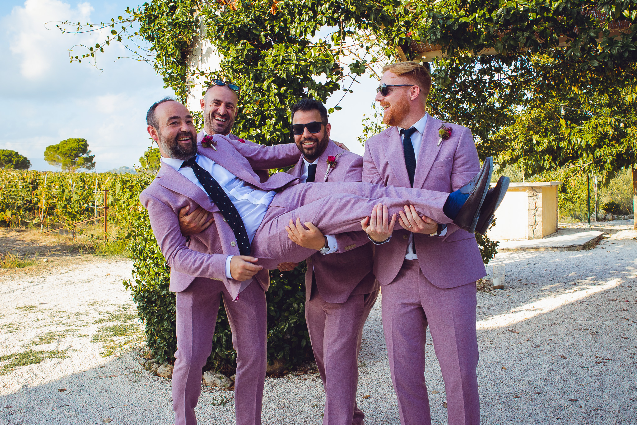 Three groomsmen laugh as they hold up the groom whilst posing for a group shot in Ambelonas Vineyard, Corfu