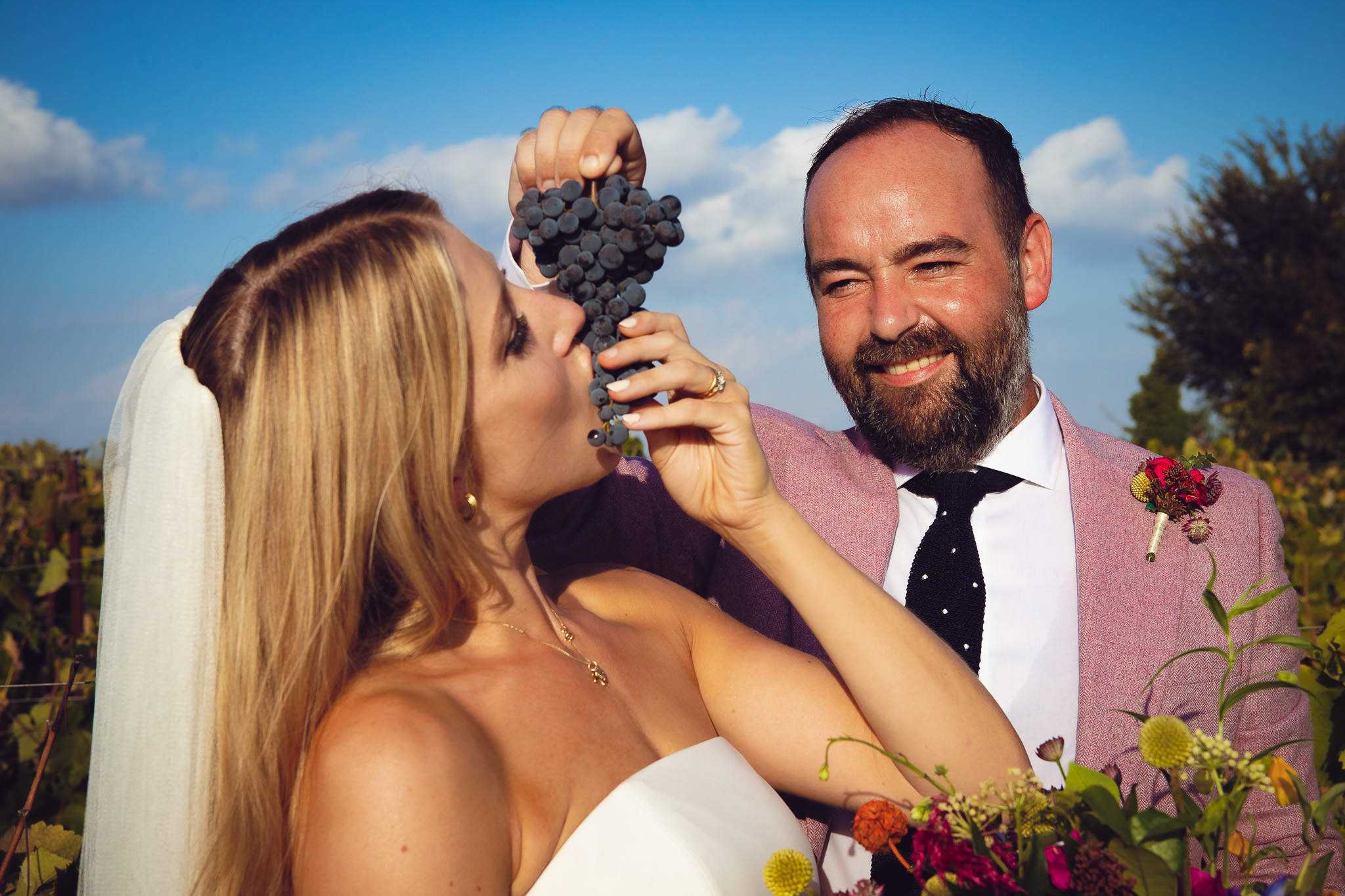 Tom holds up a bunch of grapes and feeds Alice in the vineyard during their couples photo session