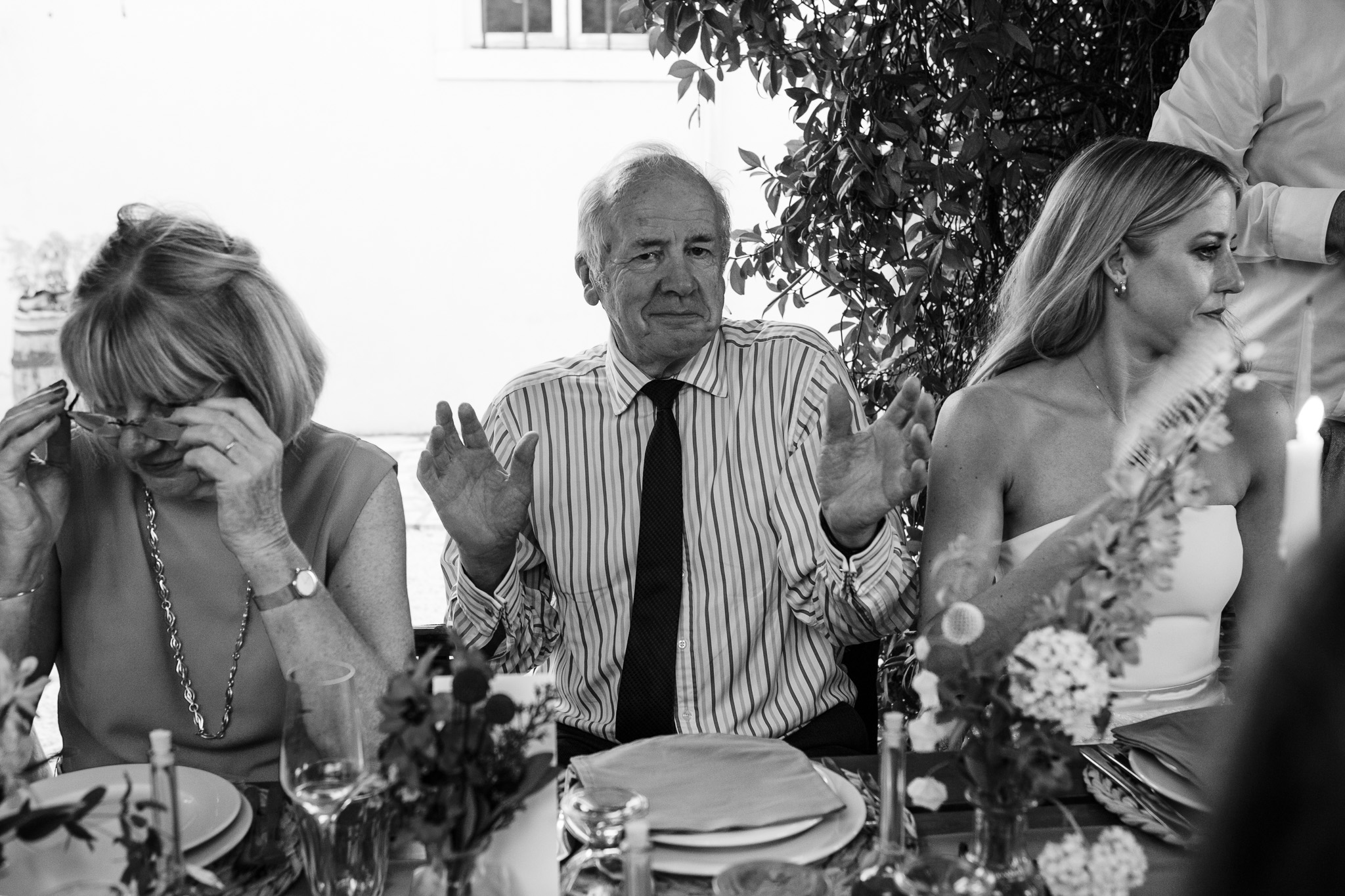 Father of the bride raises his hands as he sits between the bride and his wife at a wedding dinner