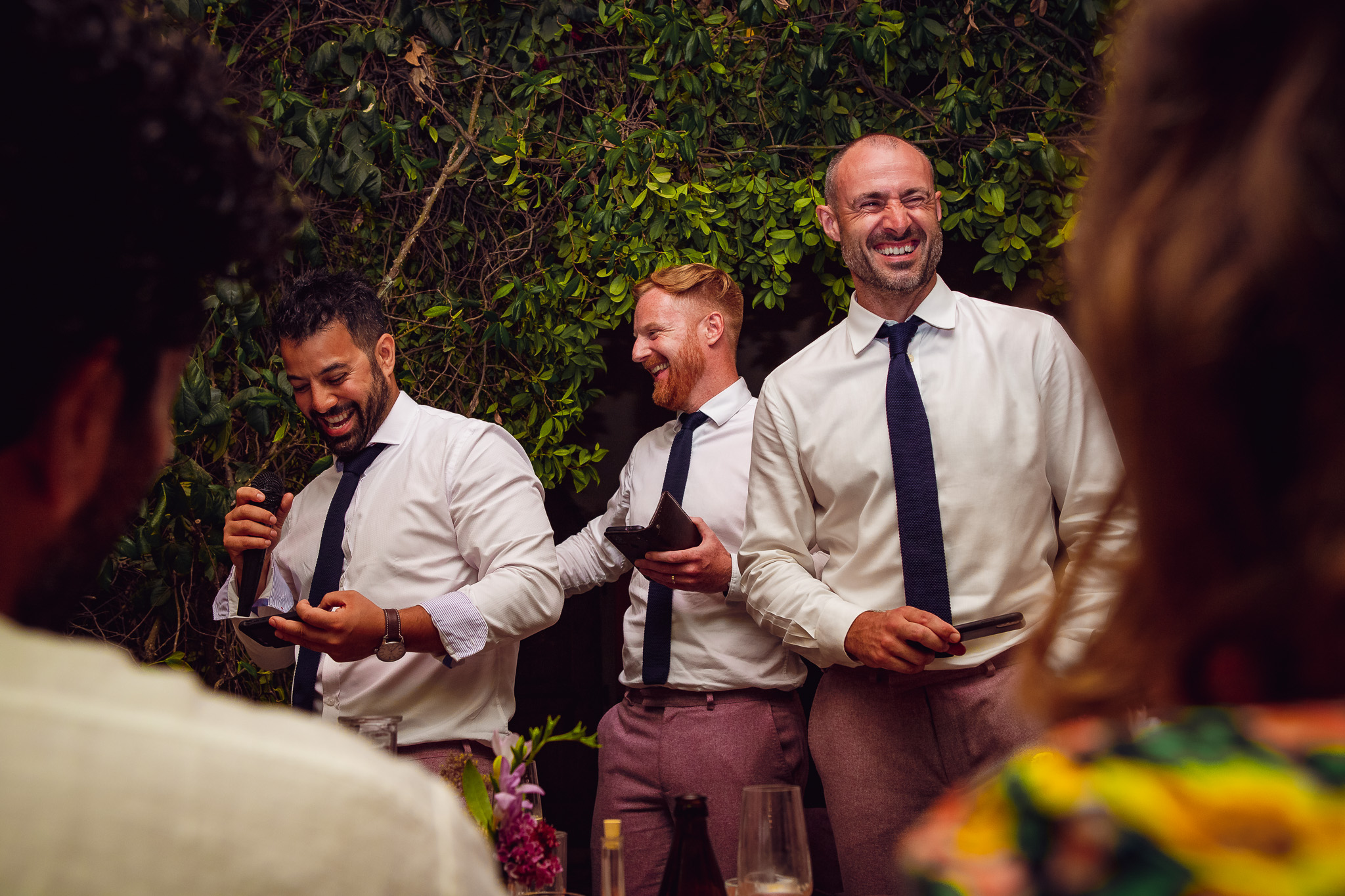 Three best men laugh as they stand to deliver their speech during a wedding dinner