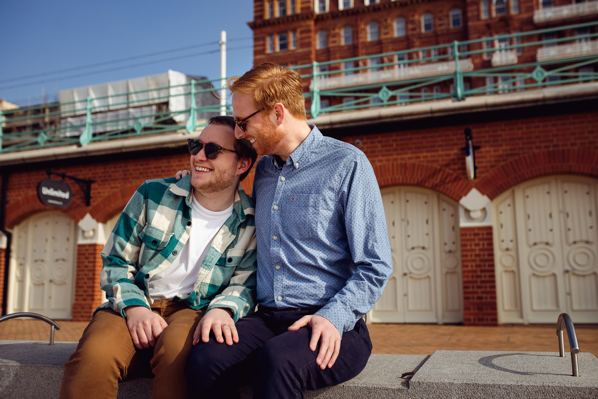 LGBTQ+ couple smile and sit on a bench on Brighton beach promenade at their engagement session