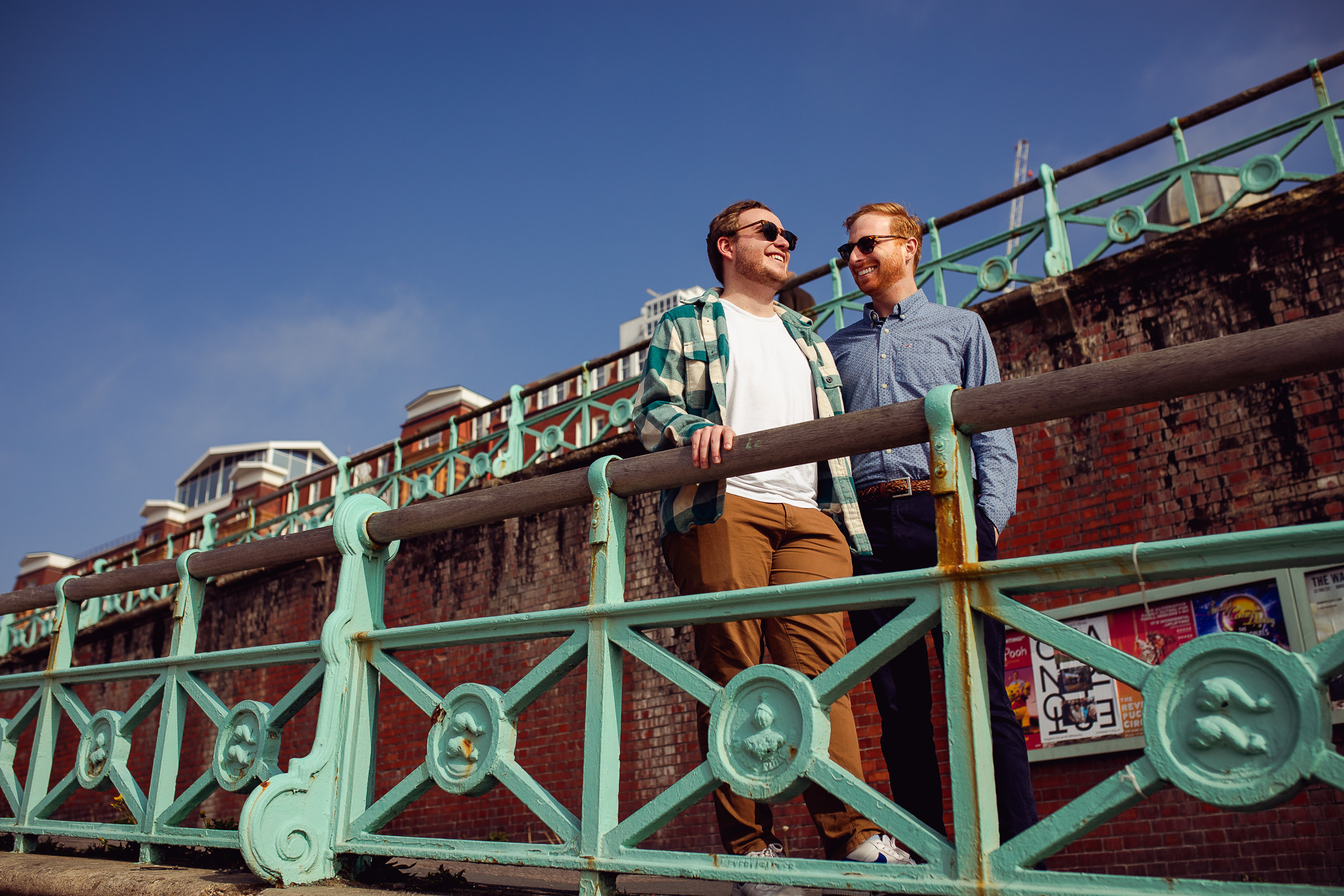 LGBTQ+ couple stand and smile on the ramp down to Brighton Beach at their engagement session