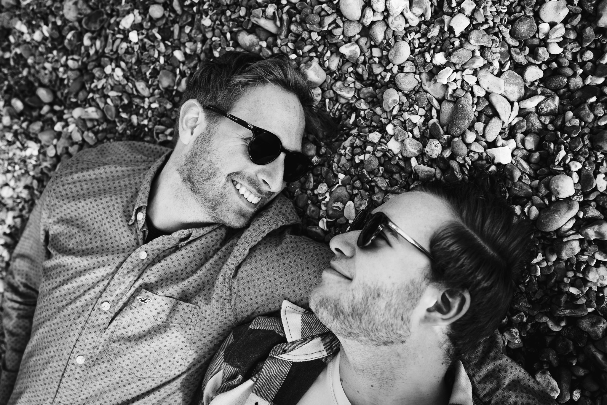 LGBTQ+ couple lying on a pebbled beach smiling and looking at each other at their engagement shoot