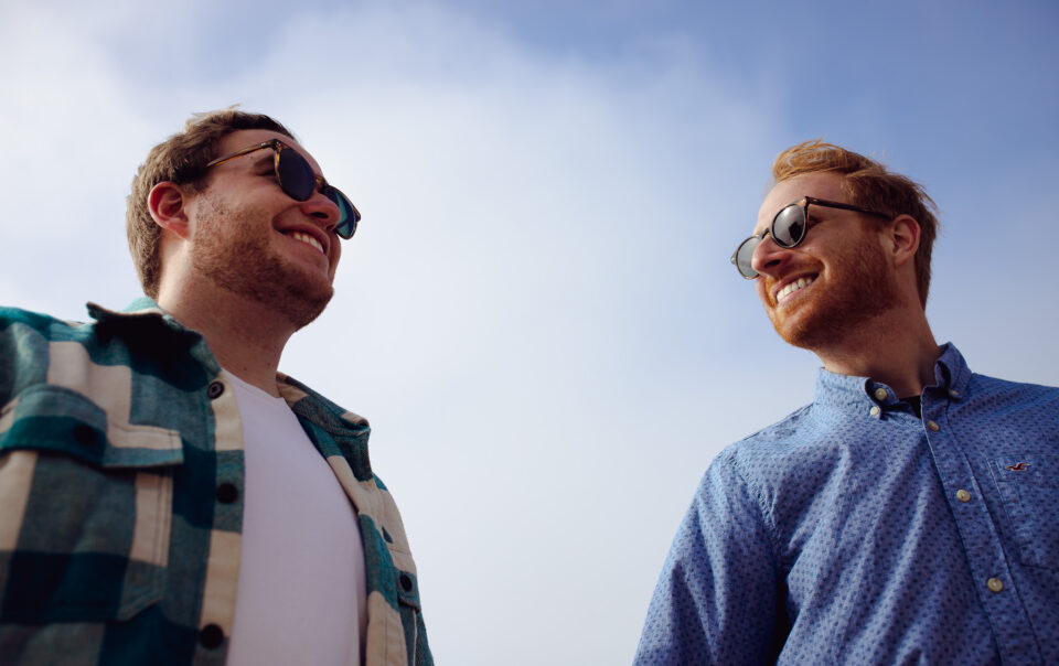 Engagement portrait of an LGBTQ+ couple wearing sunglasses and smiling at each other