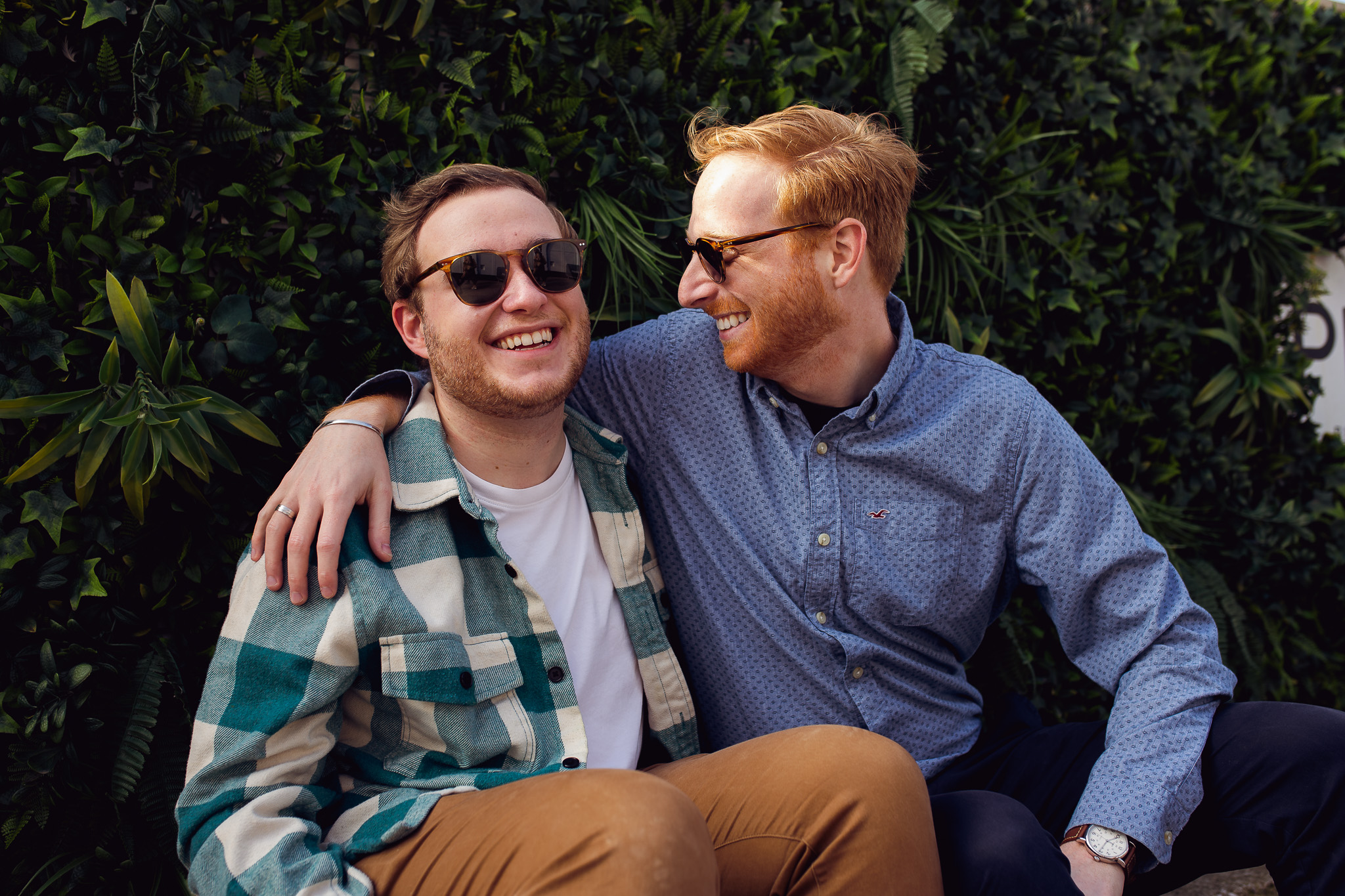 Engagement portrait of an LGBTQ+ couple wearing sunglasses and smiling sitting in front of a bush