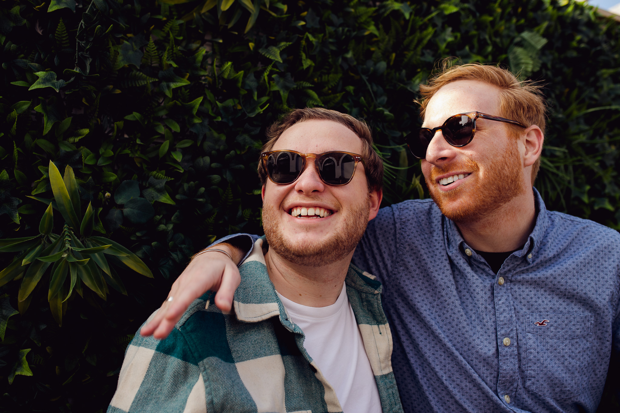 LGBTQ+ couple during their engagement session wearing sunglasses and smiling next to a bush