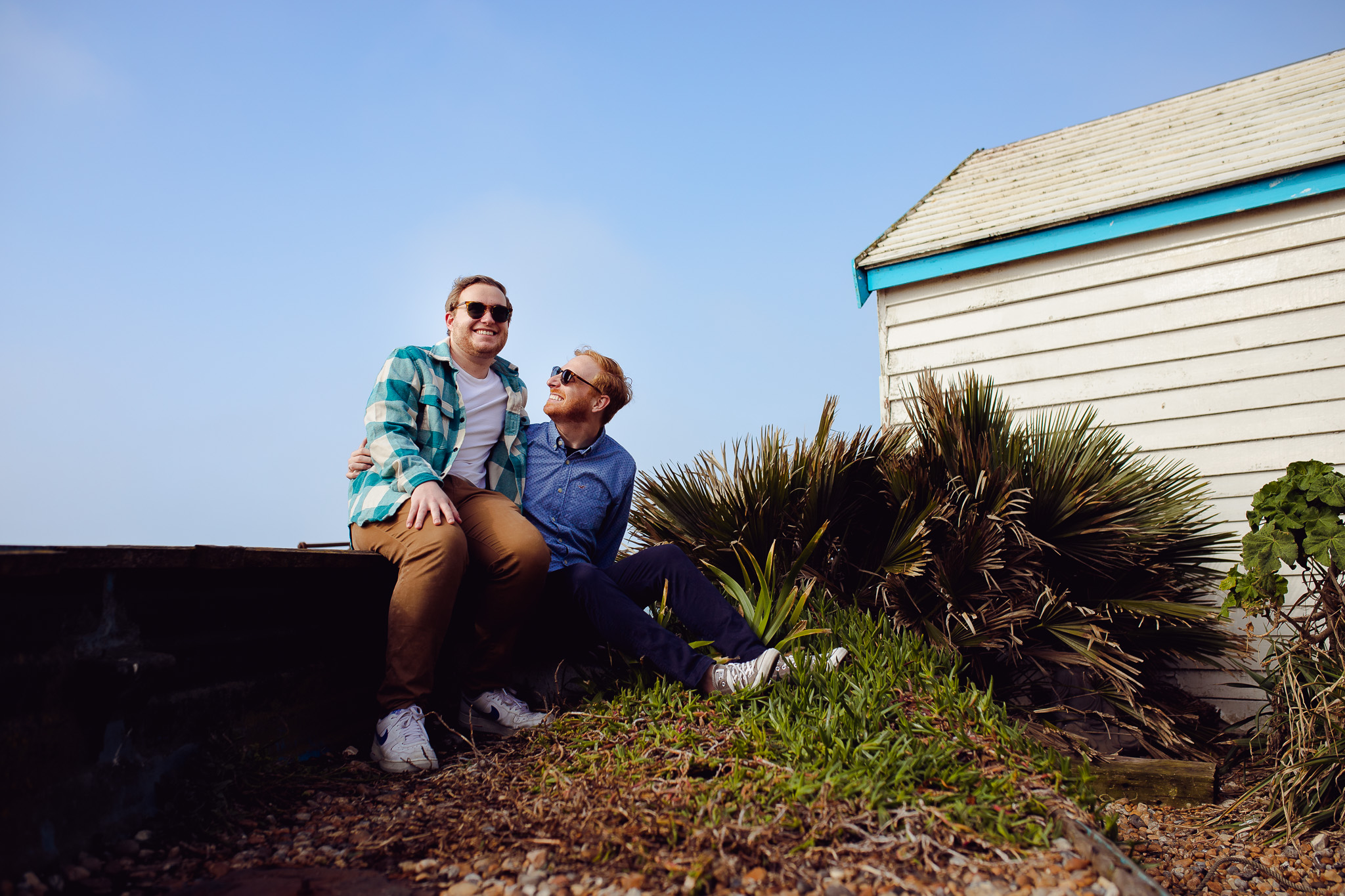 Engagement portrait of an LGBTQ+ couple wearing sunglasses and smiling sitting next to a beach hut