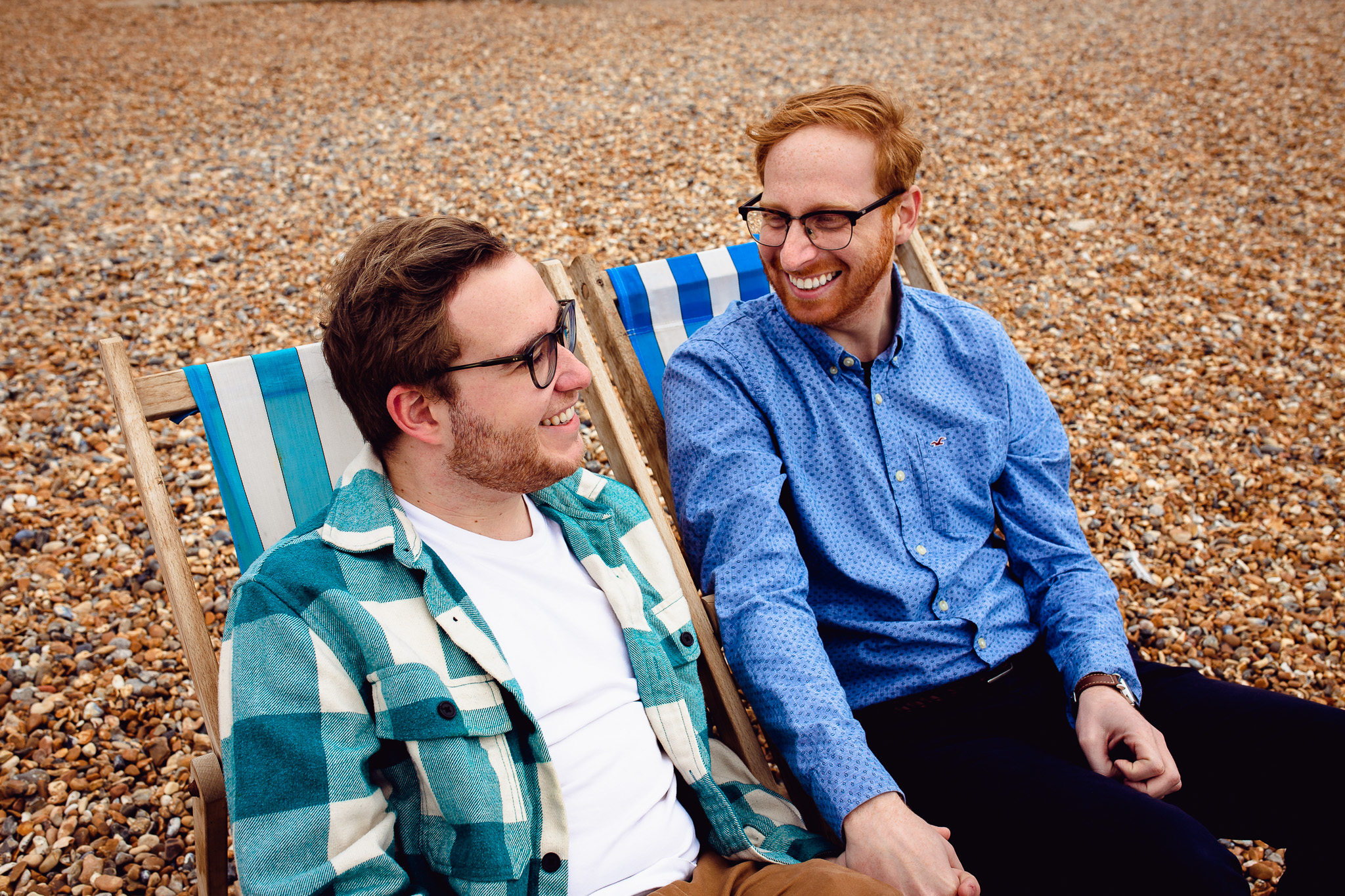 LGBTQ+ couple smiling and holding hands and sitting on deck chairs during their engagement session