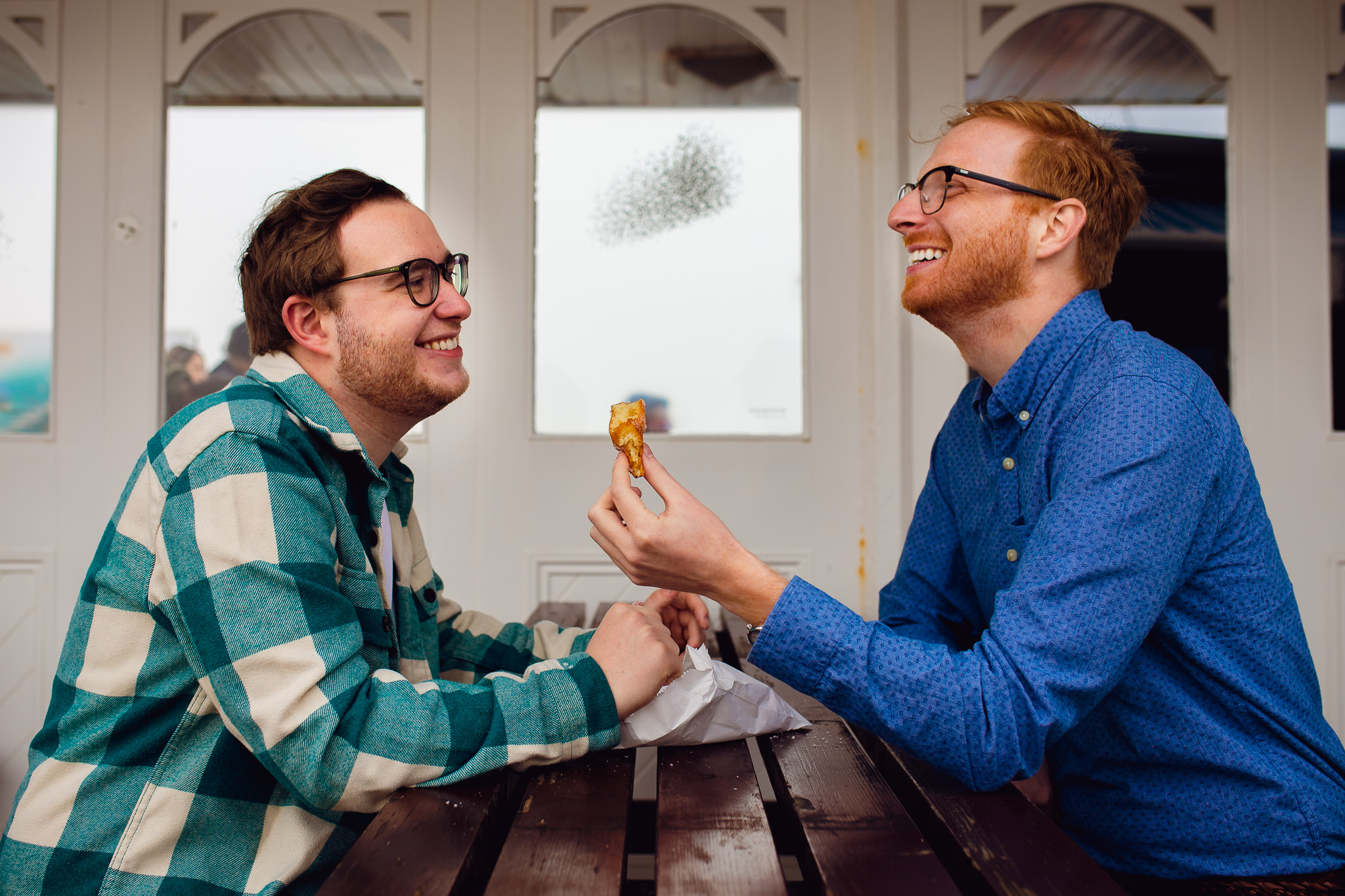 LGBTQ+ couple sitting at a picnic table laughing and eating donuts during their engagement session