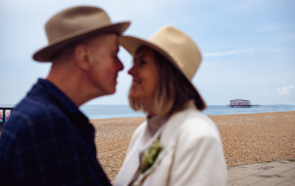 Mike and Mary touch fedoras as they look at each other during their couple portrait session with Brighton West Pier in the background.