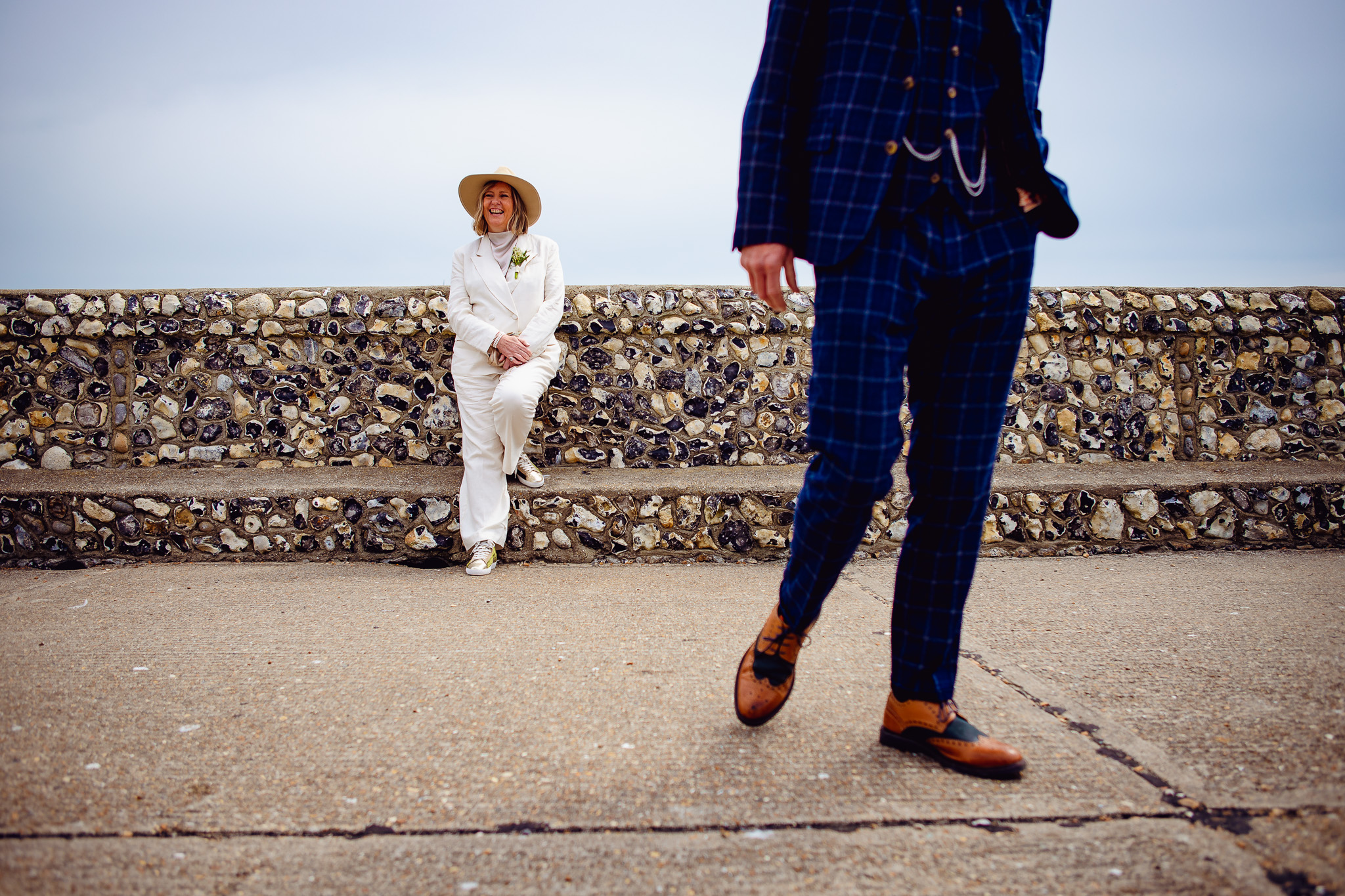 Mary stands and laughs in front of a stone wall near Brighton pier whilst Mike walks towards the photographer.