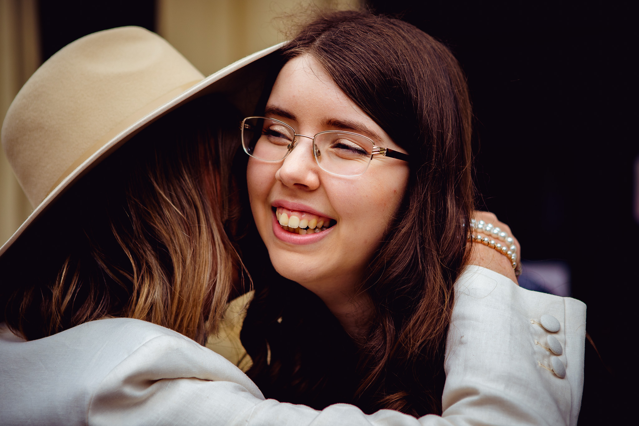 The girl smiles as she is hugged and greeted by the bride who's wearing a fedora.