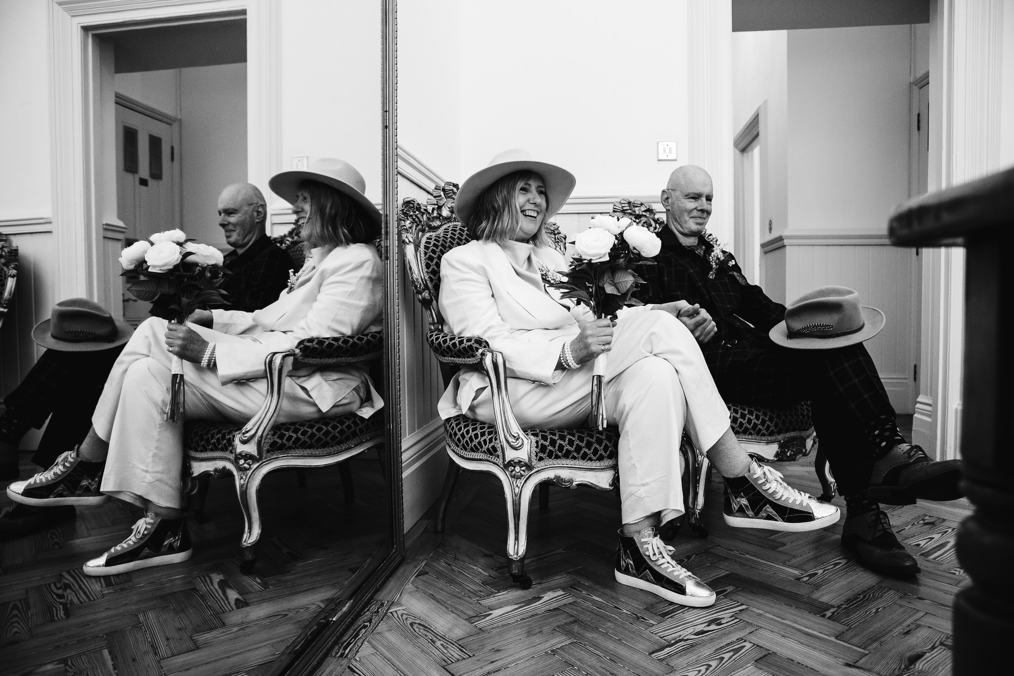 Mary and Mike sit together next to a large mirror holding hands and smiling waiting for their wedding ceremony to start.