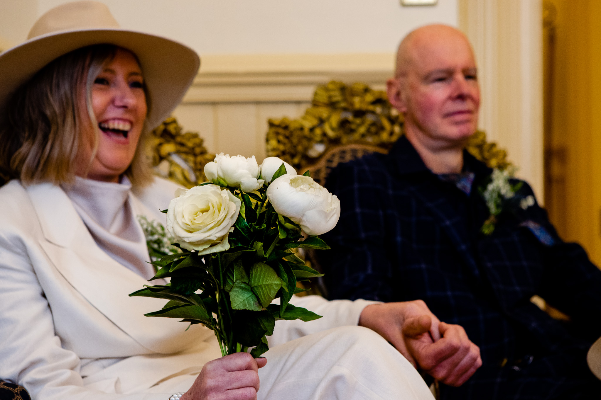 Mary holding a white bouquet and holds Mike's hand whilst sitting down at Brighton Town Hall.