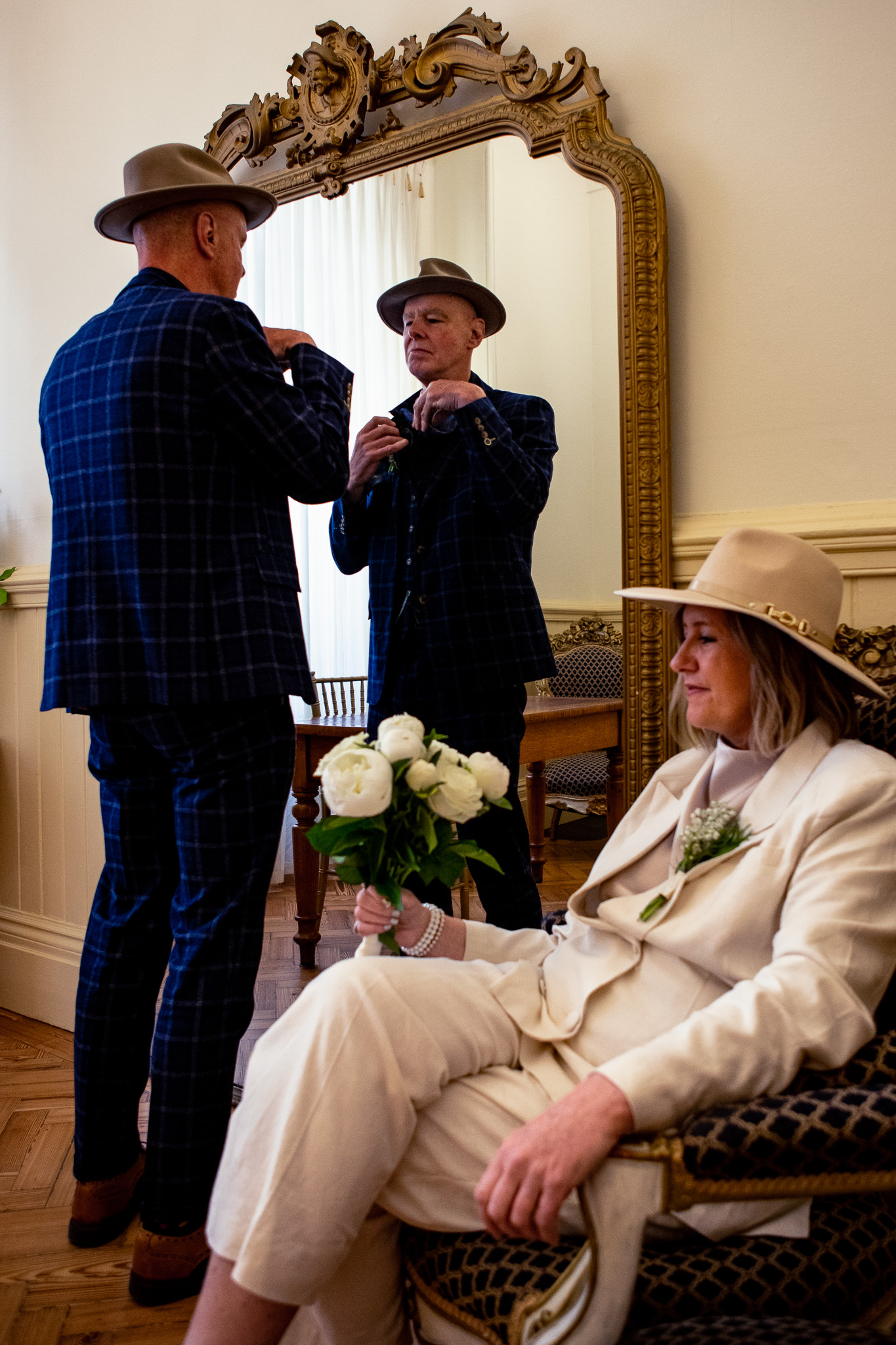 Mike adjusts his wedding outfit in the mirror whilst Mary sits next to him holding her bouquet at Brighton Town Hall.