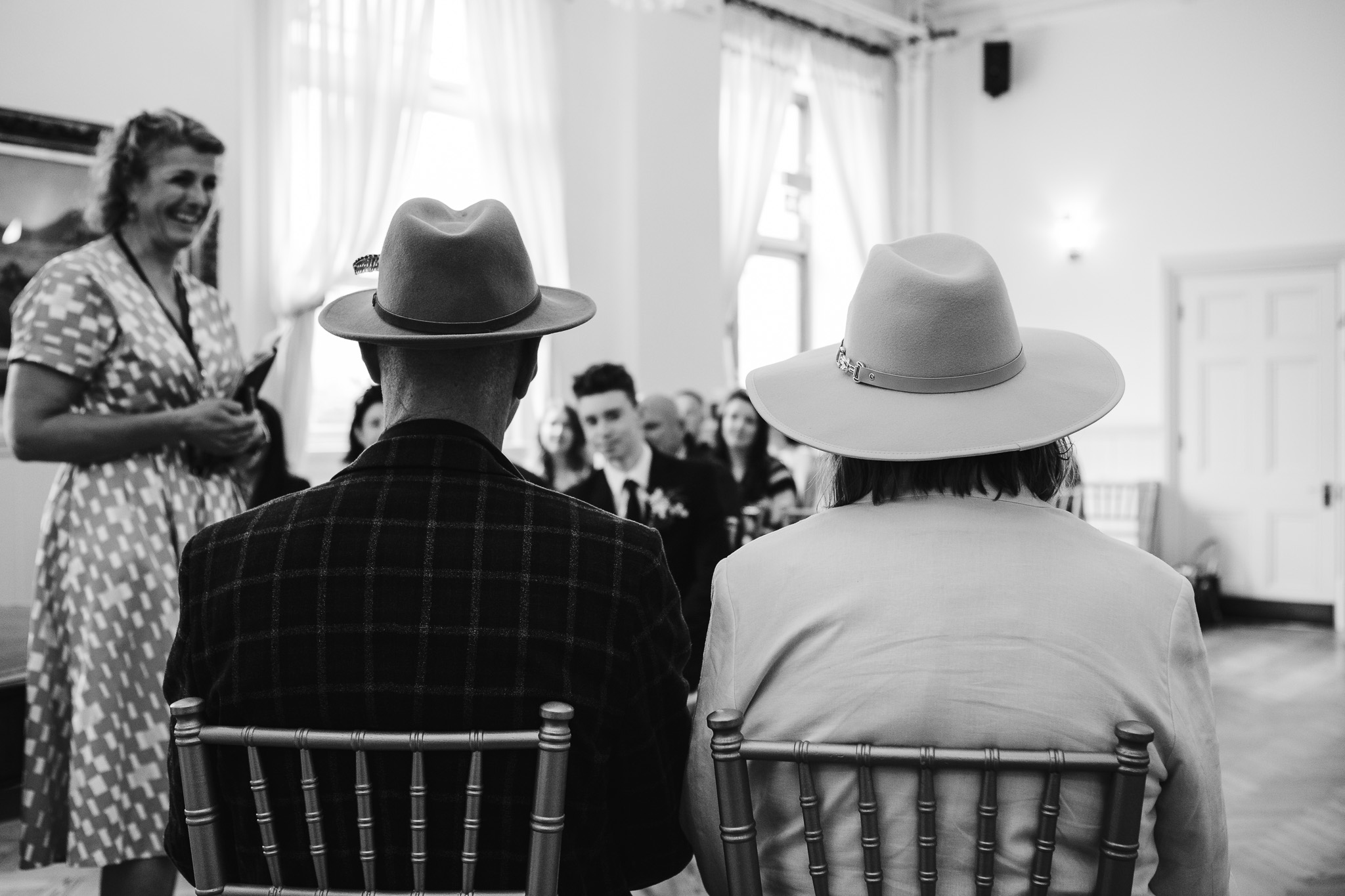 The backs of Mary and Mike wearing their fedoras sitting in front of their guests at their wedding ceremony at Brighton Town Hall.