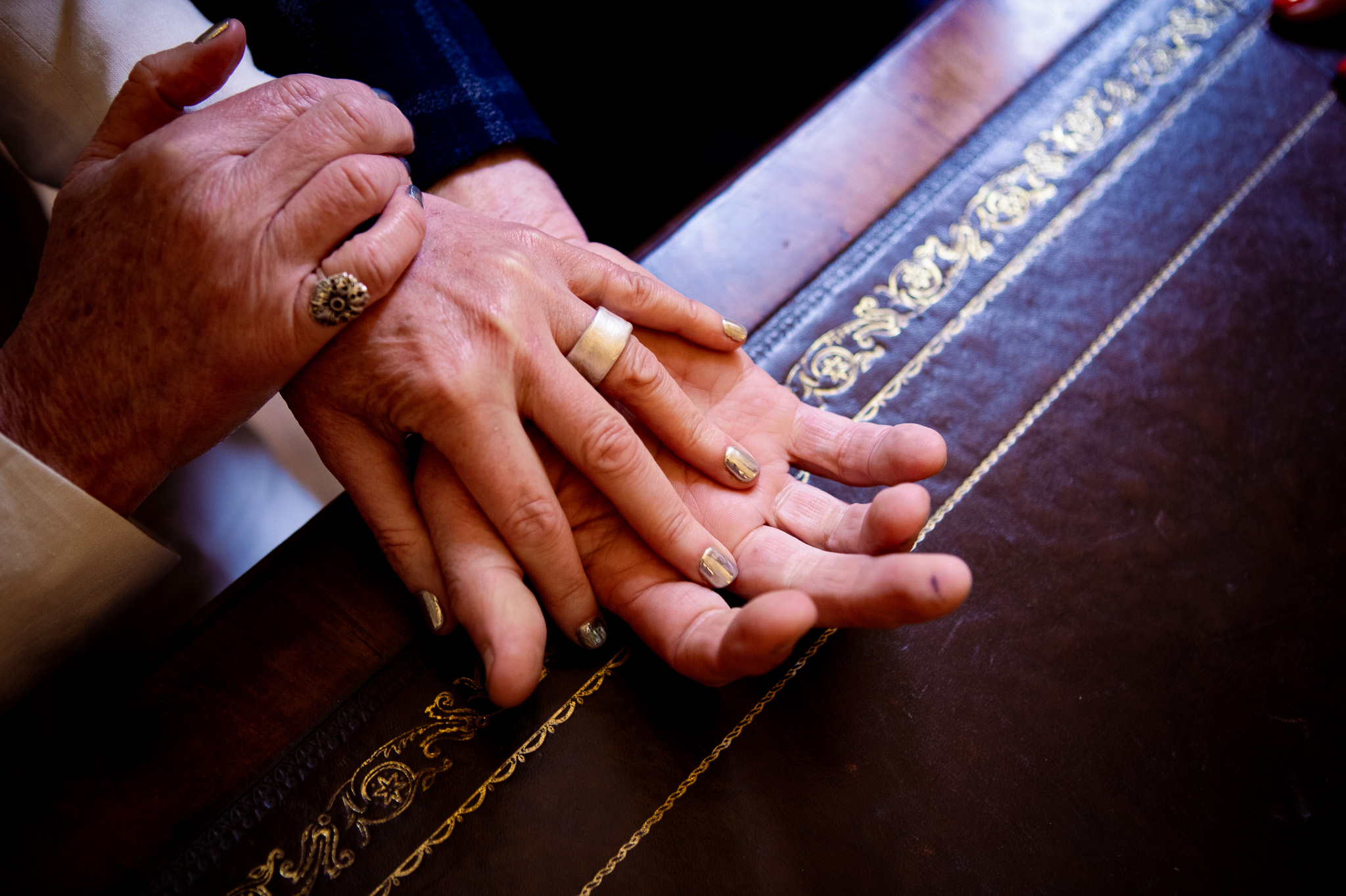 The groom holds out his hand on a desk with his newly wed wife's hand in his wearing her wedding band.
