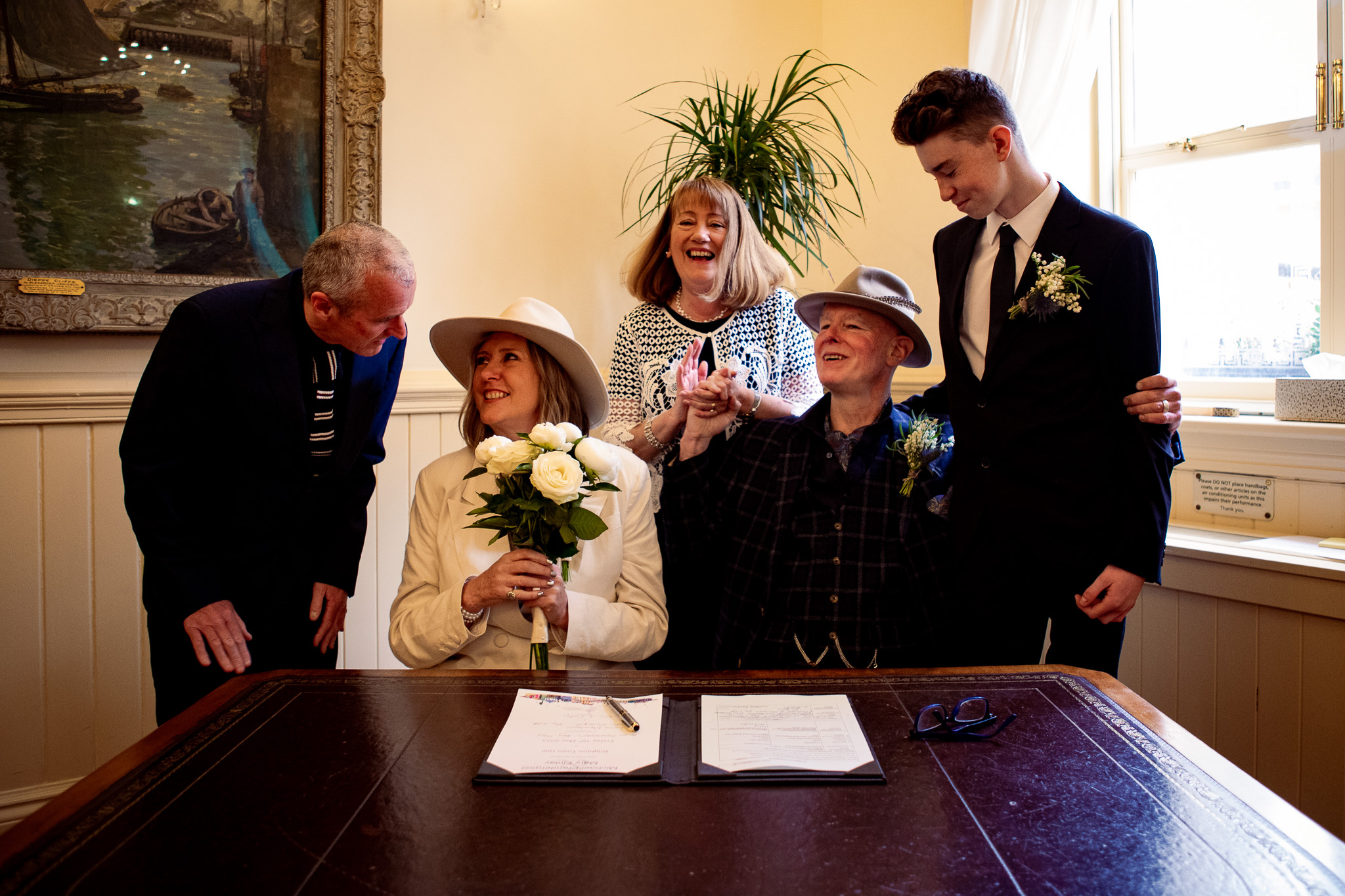 Mike and Mary sit laughing at a desk with their marriage certificates with their two witnesses and their son at Brighton Town Hall.
