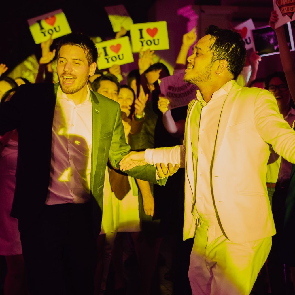 LGBTQ+ dancing at their wedding reception surrounded by guests holding I love you placards.