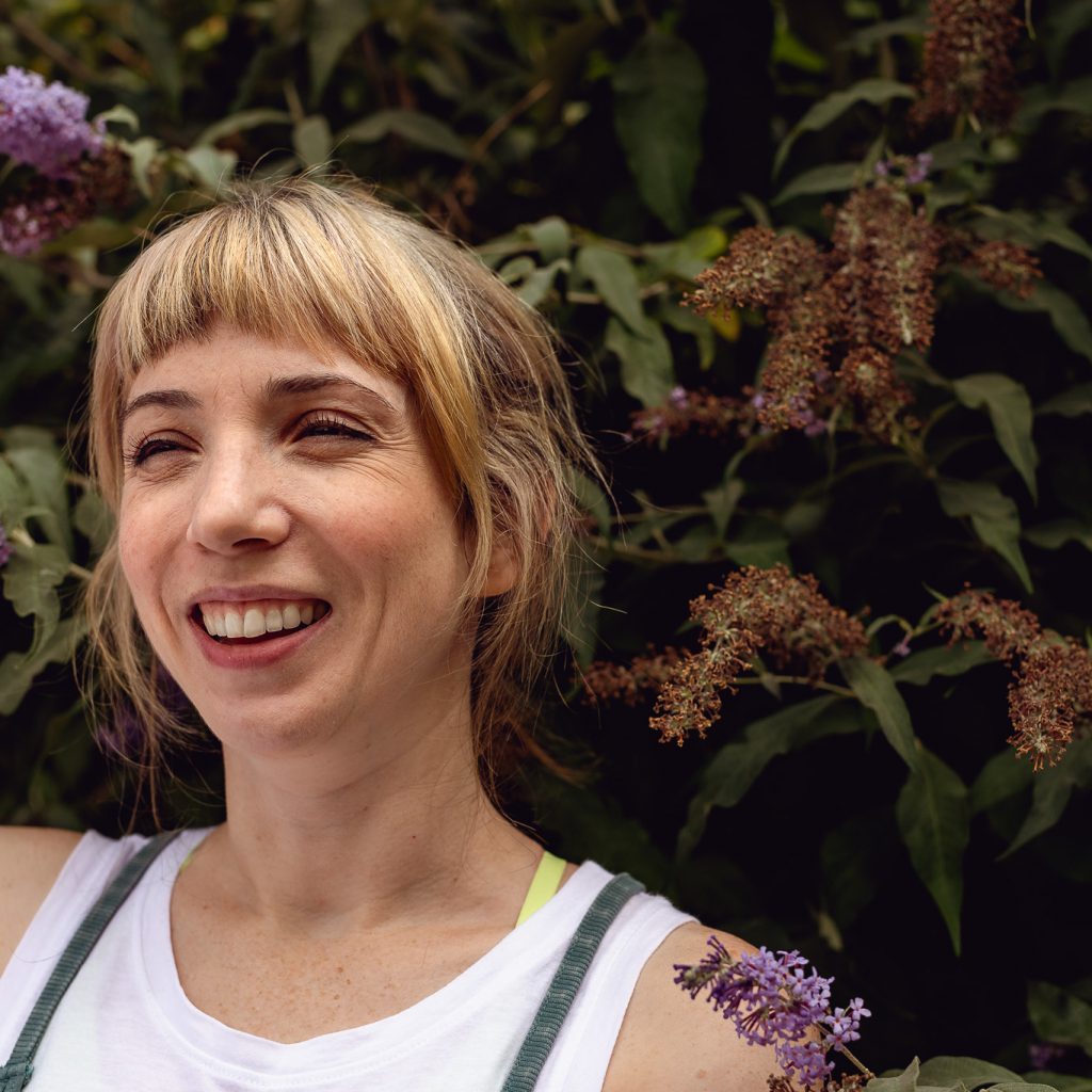 outdoor headshot of a smiling young woman standing in front of a bush with purple flowers