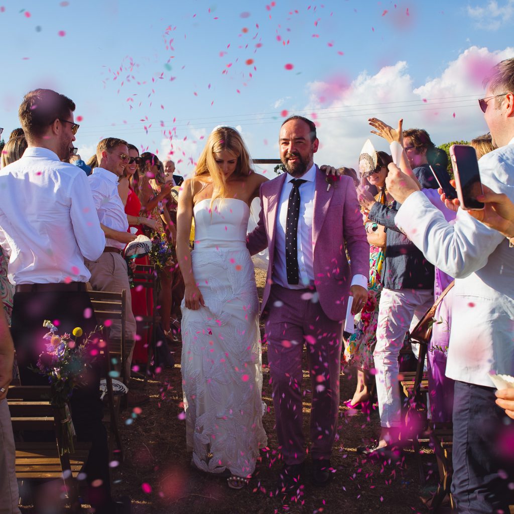 The bride and groom walk through their guests and pink confetti at their wedding ceremony